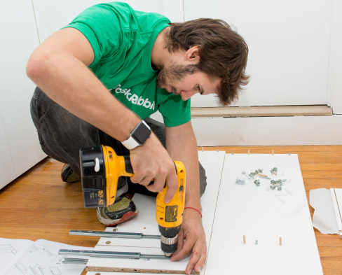 Um homem com uma parafusadeira na mão montando um móvel, parece ser um armário.